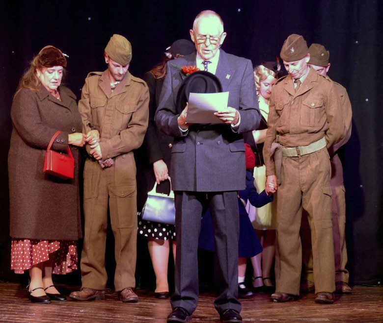 The narrator reading on stage among the cast